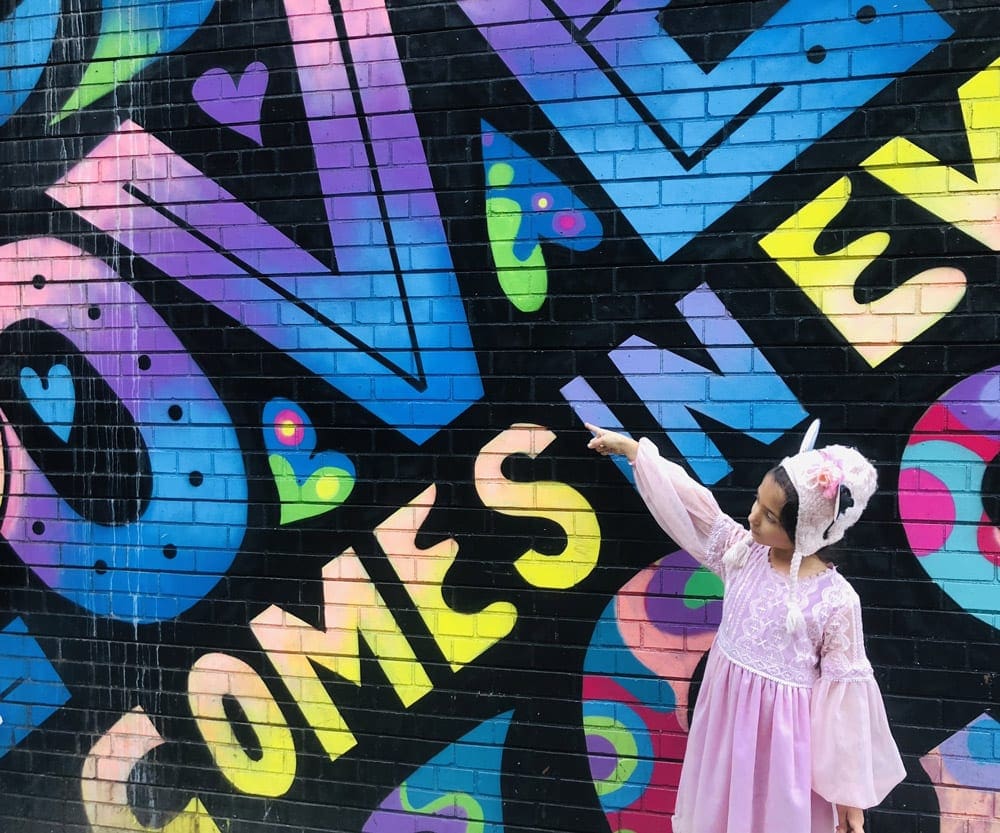 A young girl, dressed all in pink, points to a vibrant mural on a building featuring hues of blue, purple, green, and yellow.
