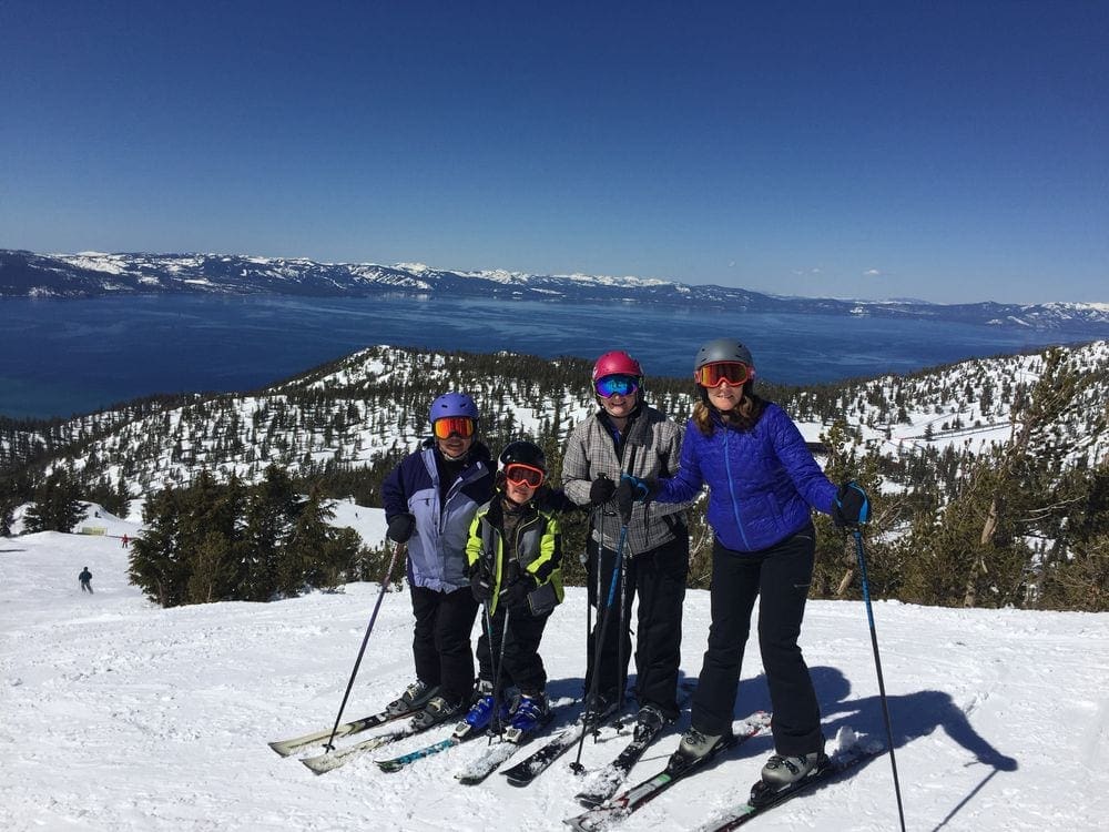 Three adults and a young boy stand on skis in full winter gear at the top of a ski hill near Lake Tahoe.