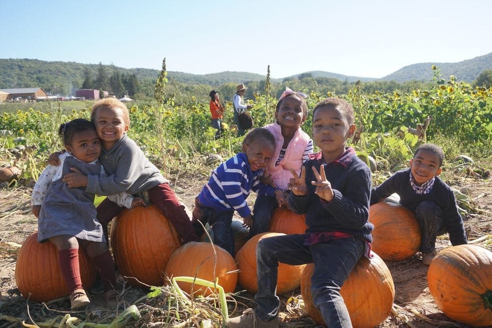 Six black kids sit on several large pumpkins, all with huge smiles, at Fishkill Farms.