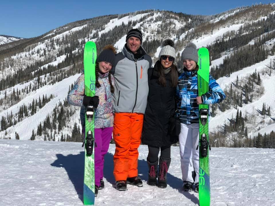 A family of four wearing snow gear holds up two large green skis at Steamboat, one of the best Colorado ski resorts for families, with a mountainous view behind them.