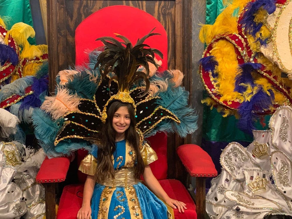 A young girl wearing Mardi Gras attire sits in a huge, plush chair showing off her colorful costume. One of the best things to do in New Orleans with kids