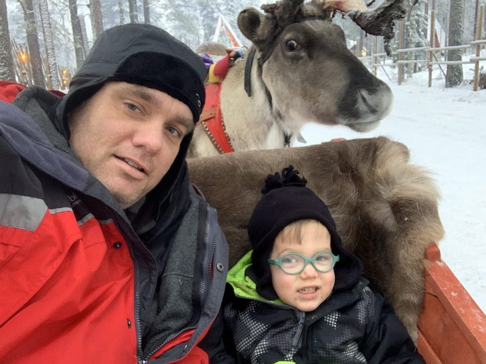 A dad and his young son pose while sitting on a sled, with a reindeer behind them.