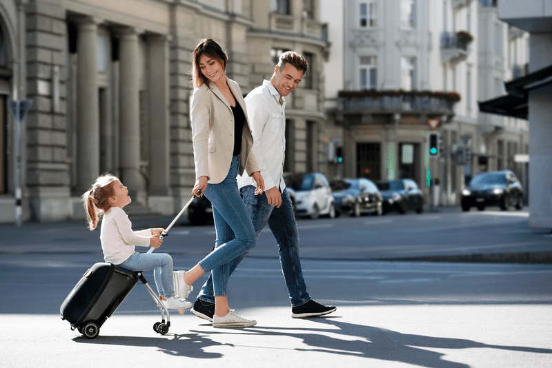Little girl rides happily on a black Micro Kickboard luggage while her parents pull her along the grey city streets. Micro Kickboard offers families some of the best travel luggage for kids. 