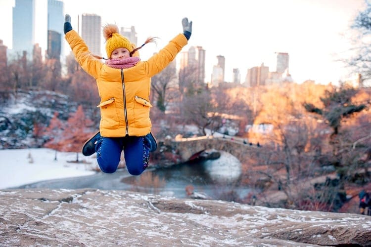 A young girl jumps for joy in Central Park, with the NYC skyline behind her.