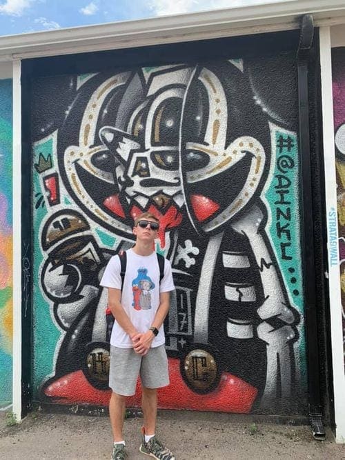 A teenager with sunglasses stands in front of street art in Kansas City, MO, featuring a robotic, skeletel image of Mickey Mouse.