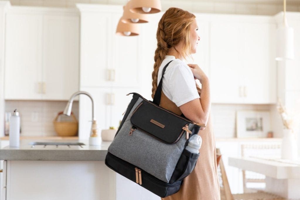 red-haired woman standing in kitchen with a black and grey Petunia Pickle Bottom bag. Petunia Pickle Bottom has some of the best travel luggage for kids. 
