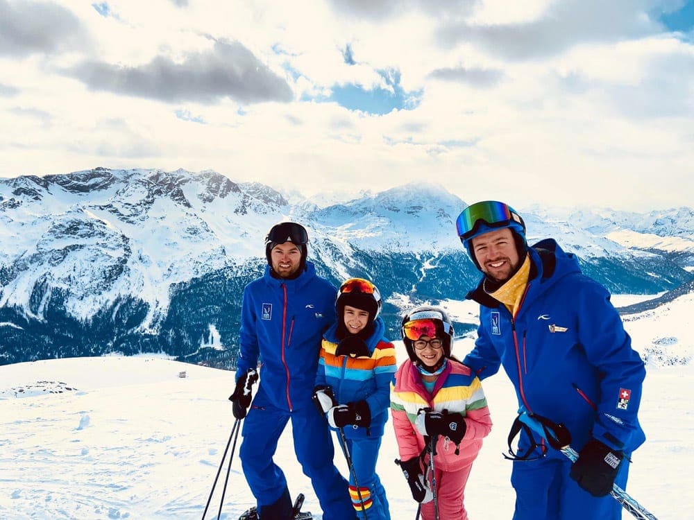 Two young children stand with their ski instructors at Suvretta House, with mountains in the background.