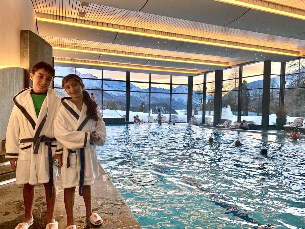 Two kids smile while wearing their Suvretta House robes and slippers, near the indoor pool, two things to remember in our Ultimate Ski Trip Packing List For Families This Winter.