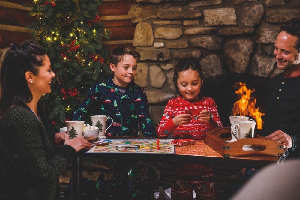 A family of four enjoys a game night with warm beverages, a fire, and Christmas tree.