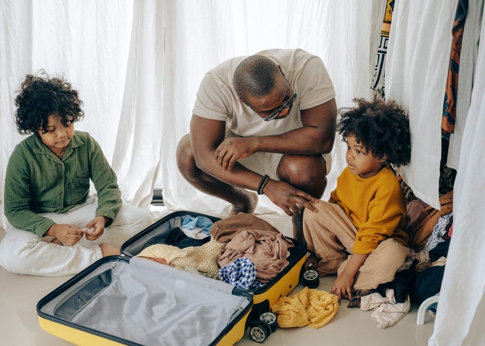 A black dad and his two kids pack a large yellow piece of luggage. Packing light is one of our tips for long car rides with toddlers