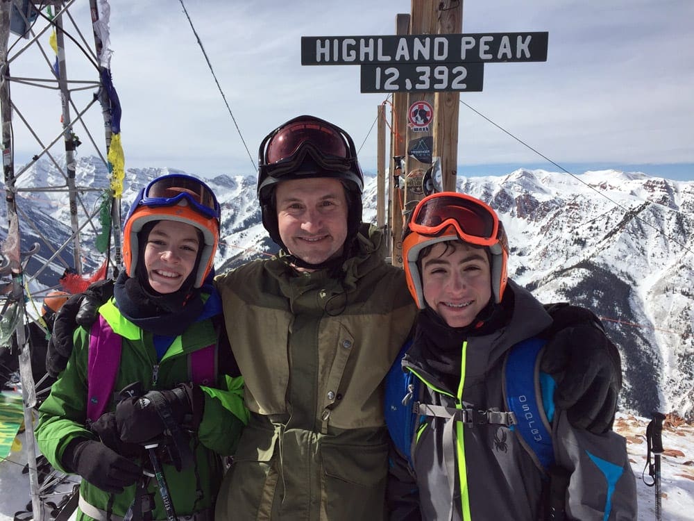 A dad and his two kids in snow gear stand in front of a sign reading "Highland Peak, 12,392" at Aspen.
