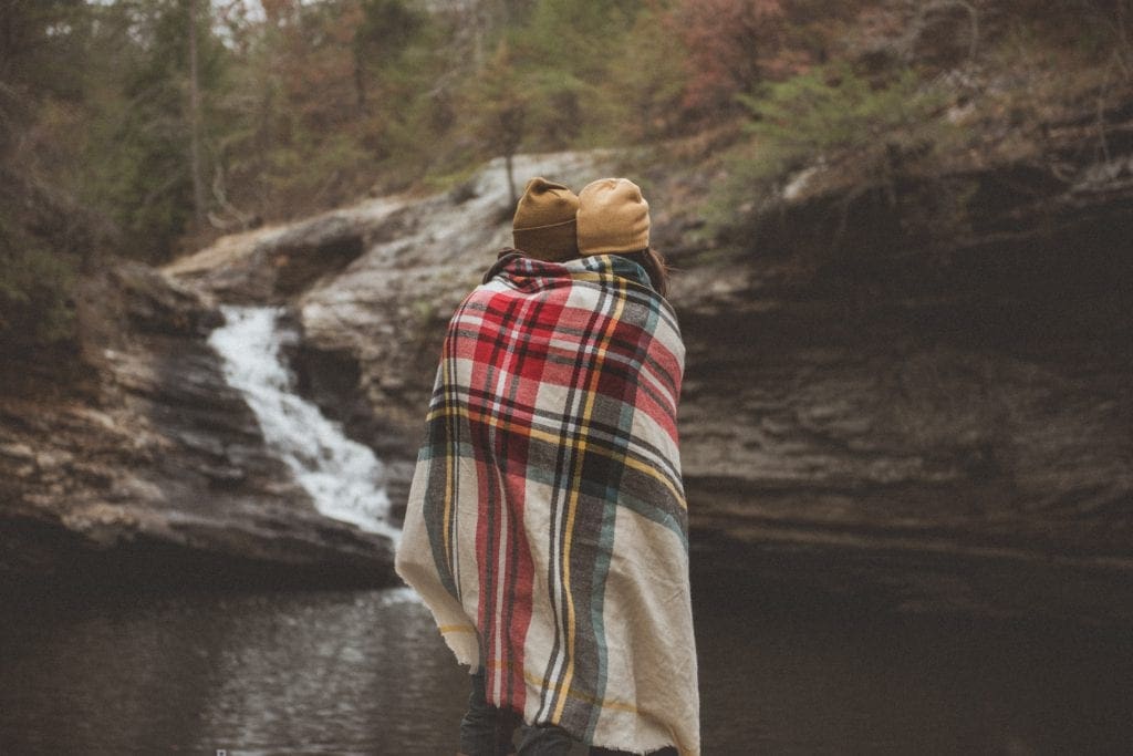 A couple is wrapped in a large blanket scarf with a waterfall and wooded area in the background.
