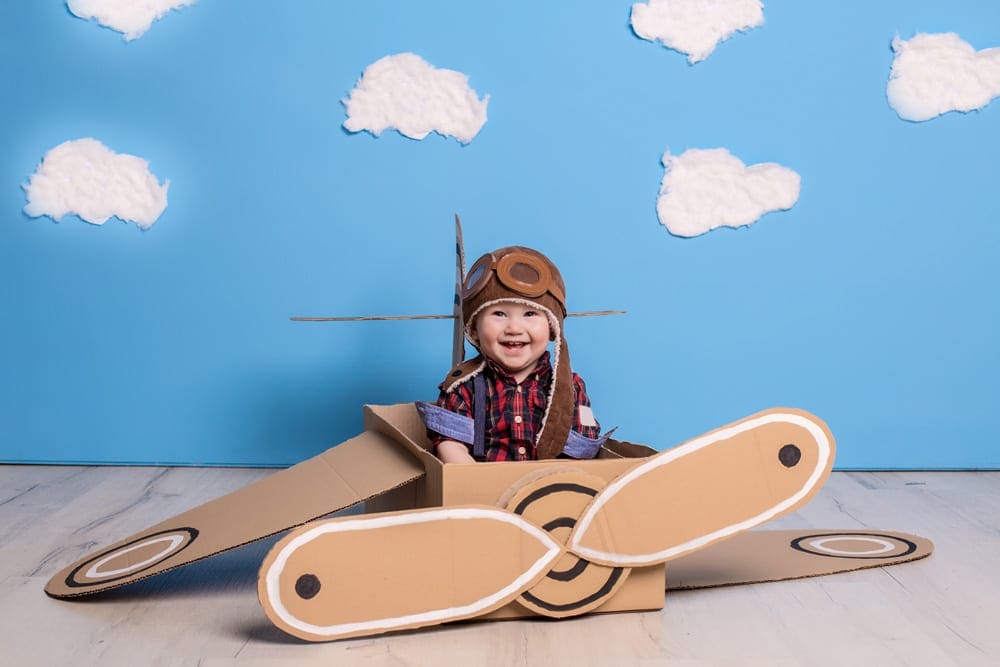 An infant wearing a pilot costume sits in a cardboard plane with a blue wall and painted clouds behind him, one of the best travel themed costumes for kids.