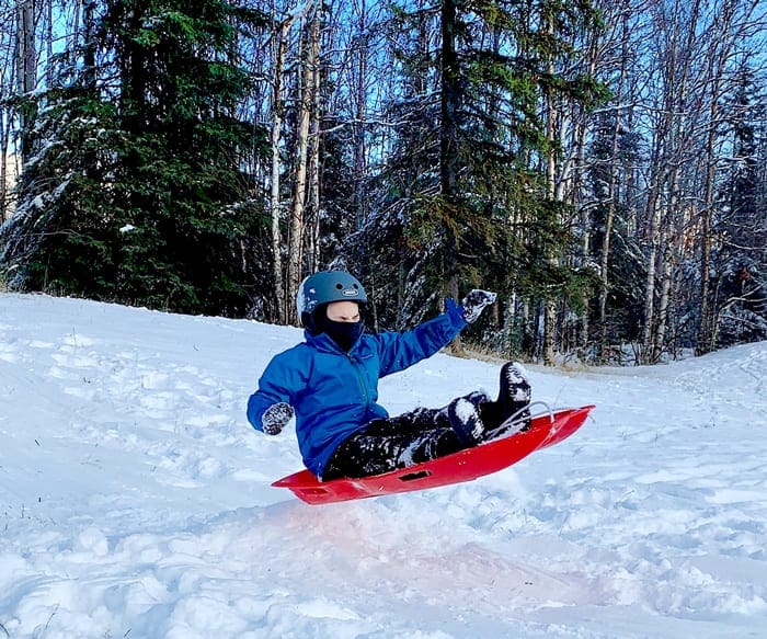 A young boy wearing a blue helmet and a blue coat sits on a red sled airborne, as it goes over a small snow mound.