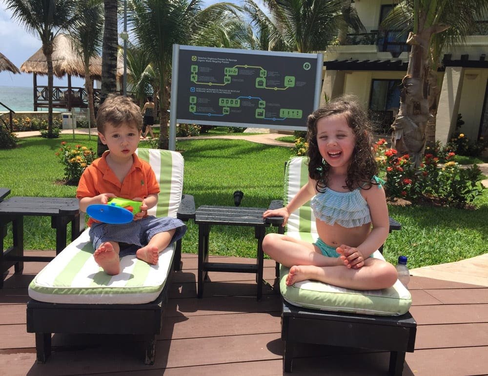Two kids sit on poolside chairs, laughing at The Fives Azul Beach Resort Playa del Carmen.