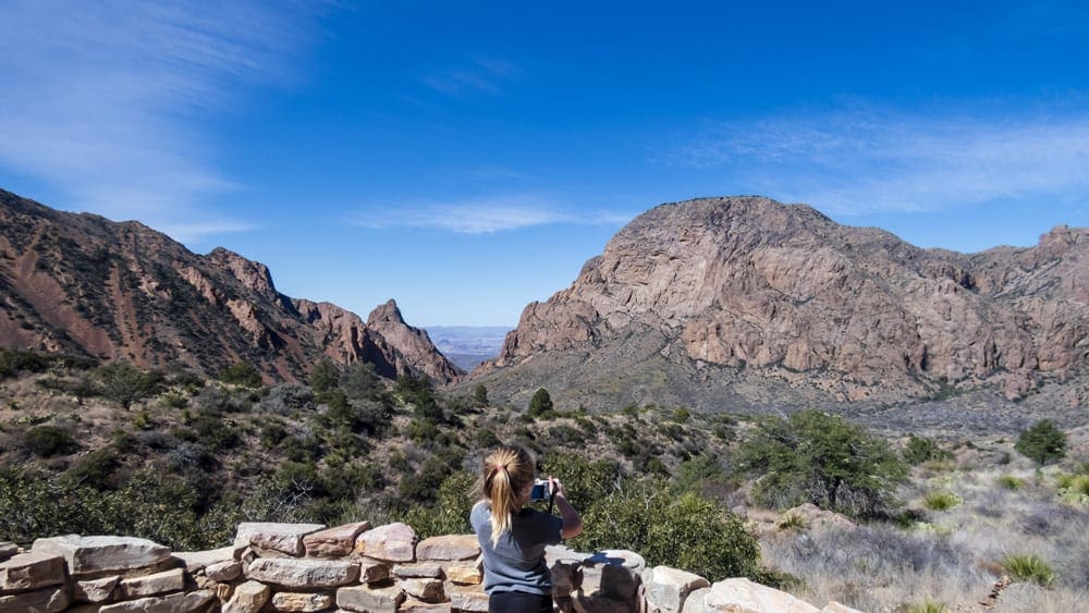 A young girl takes a picture of an expansive view of Big Bend National Park, featuring large rock mountains and sparse foliage.
