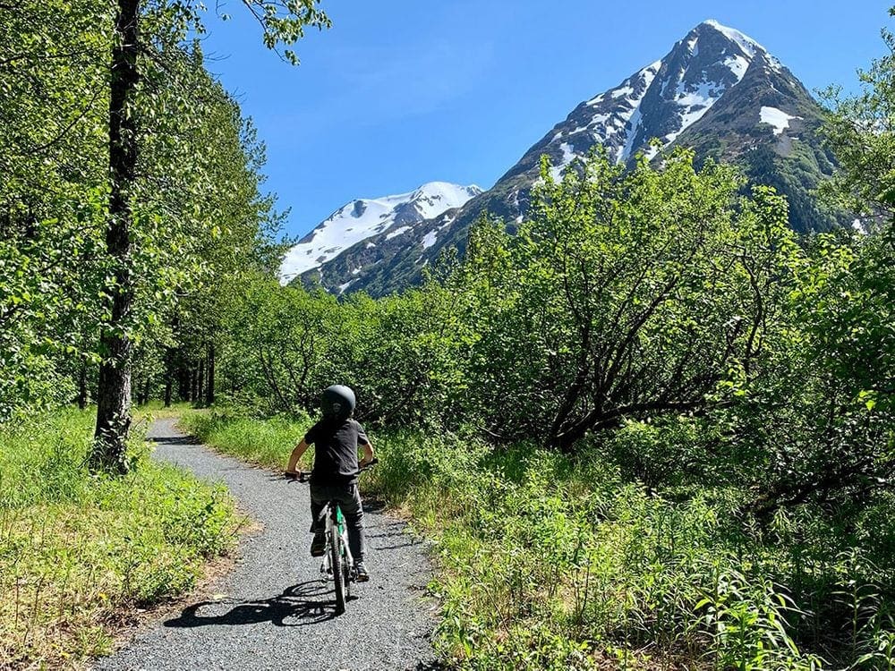 A young boy wearing a helmet bikes along a path with trees and mountains in the background. Bikes are some of the best outdoor gifts for families.