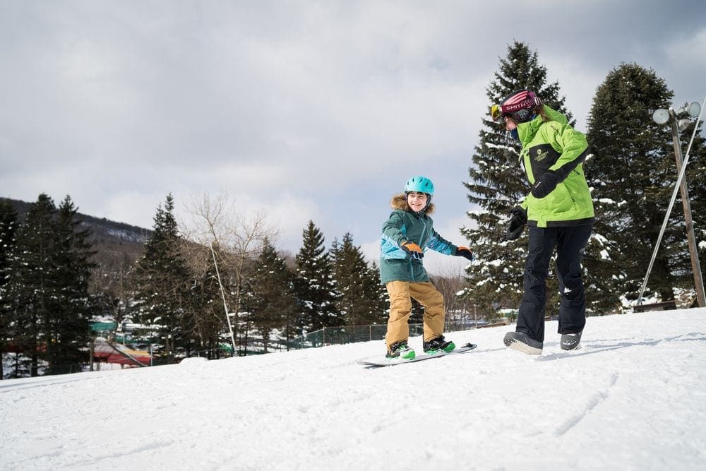 A young child slowly goes down a run on a snowboard, with their ski instructor coaching them each step of the way.