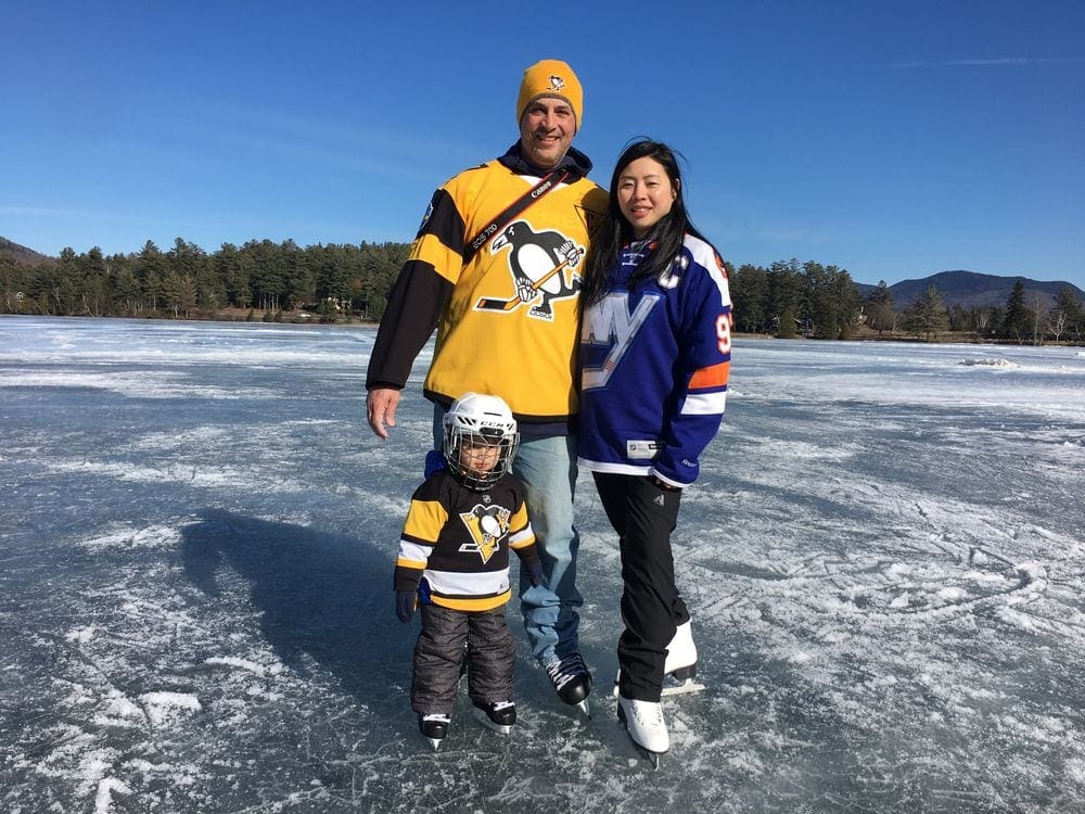 A family of three, all wearing NHL hockey jerseys, stand on an iced over Lake Placid wearing skates.