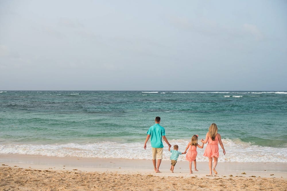 A family of four, wearing color coordinated outfits in blue and pink, walks on the beach toward the water in the Dominican Republic.