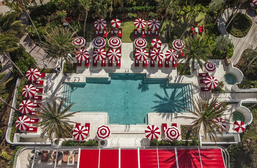 An aeiral view of a large turquois pool at Faena Hotel Miami Beach, ones of the best family-friendly hotels in Miami, surrounded by the iconic candy red chairs and umbrellas.