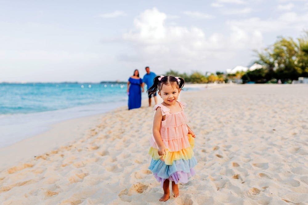 A small girl wearing a colorful dress smiles while her parents look on in the background on a beach in the Cayman Islands.