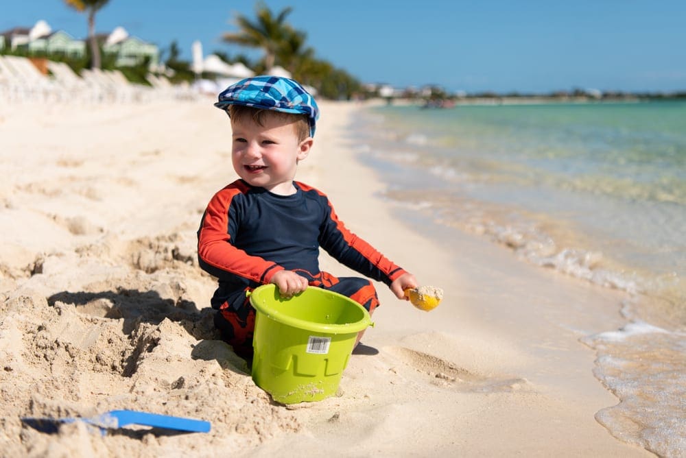 A young boy wearing a cap sits smiling in the sand on a beach.