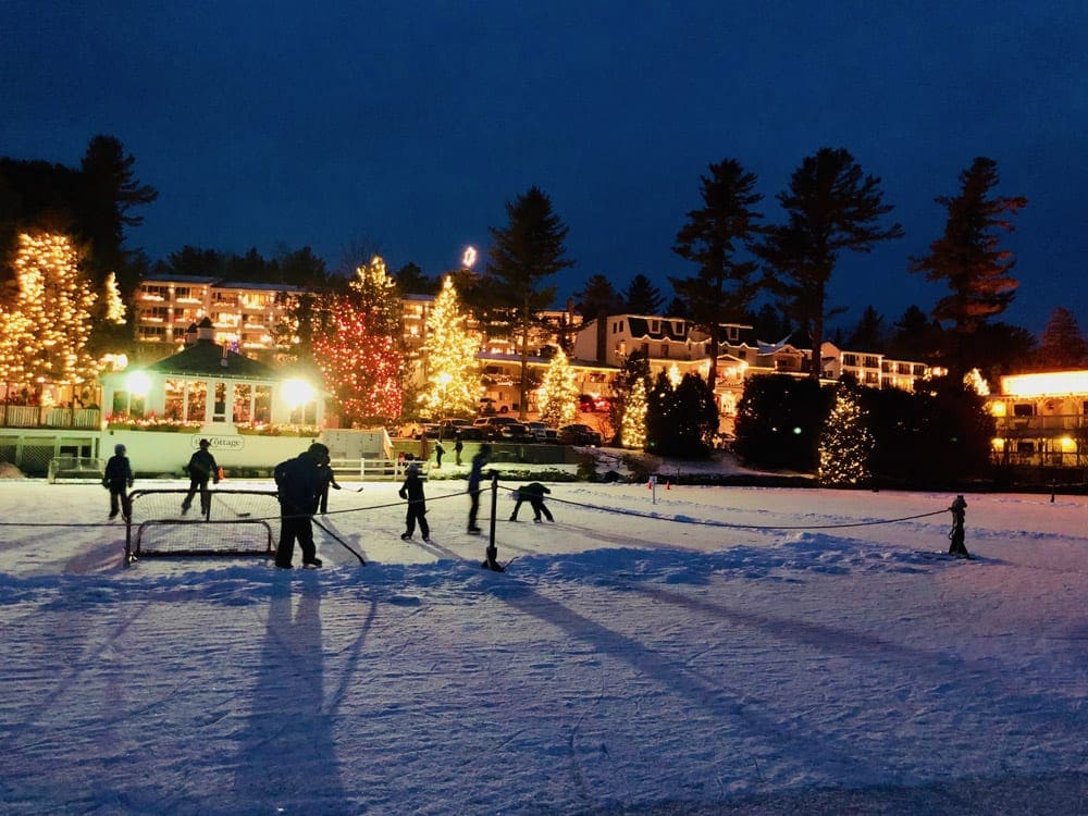Several kids play hockey on a frozen pond, while Christmas lights dot the background in Lake Placid, one of the best Christmas towns in the Northeast.