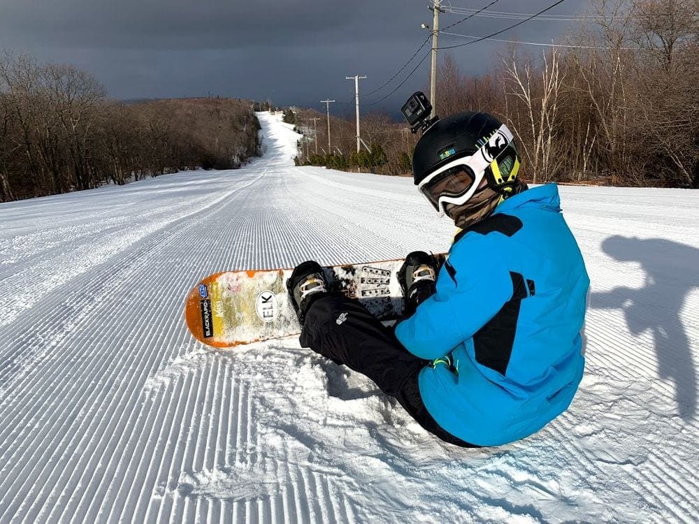 A young boy sits in the snow on a slope at Blue Mountain while resting his snowboard, one of the things to remember in our Ultimate Ski Trip Packing List For Families This Winter.