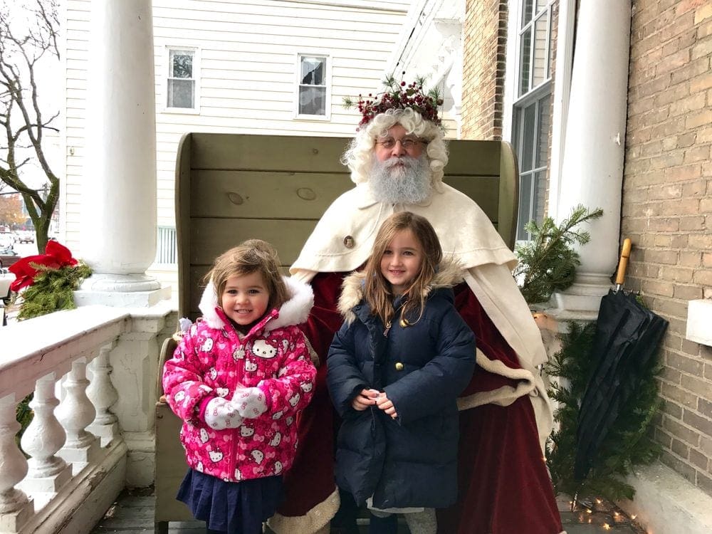 Two girls wearing large winter coats stands with Santa in Skaneateles, New York, one of the best Christmas towns in the Northeast.