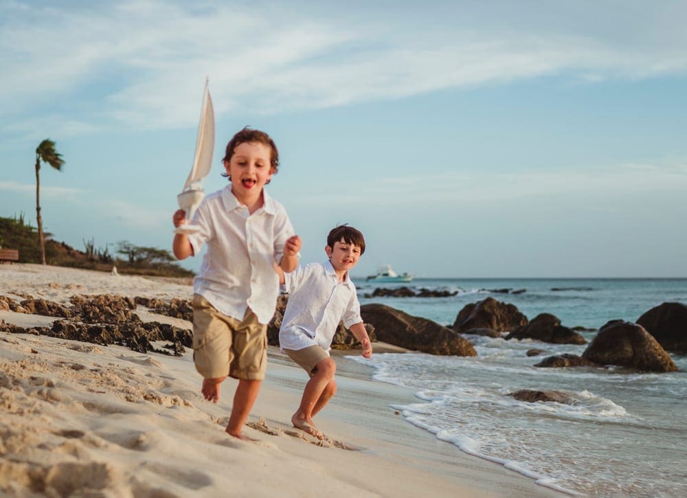 Two boys race down a beach in Aruba, while one carries a toy sail boat, one of the best hot places to visit in December with kids!