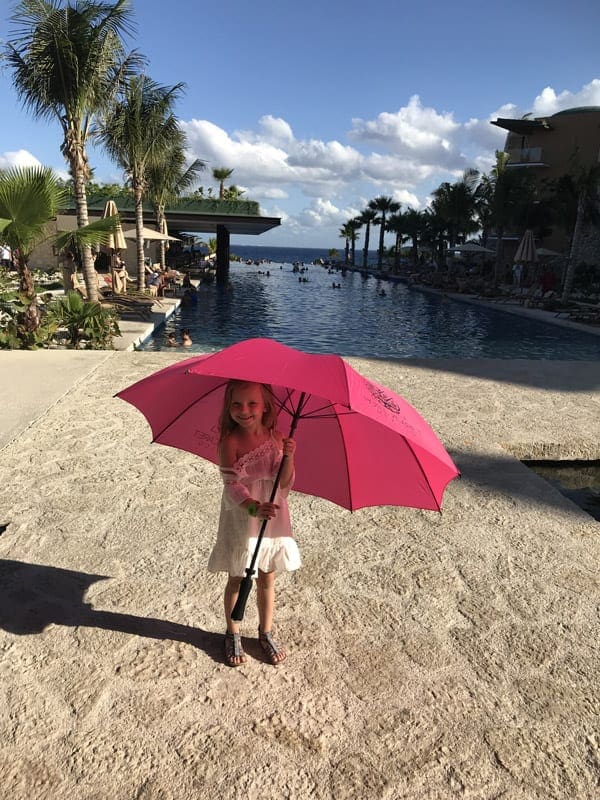 A young girl holds an opened bright pink sun umbrella with a pool behind her at Hotel Xcaret Mexico.