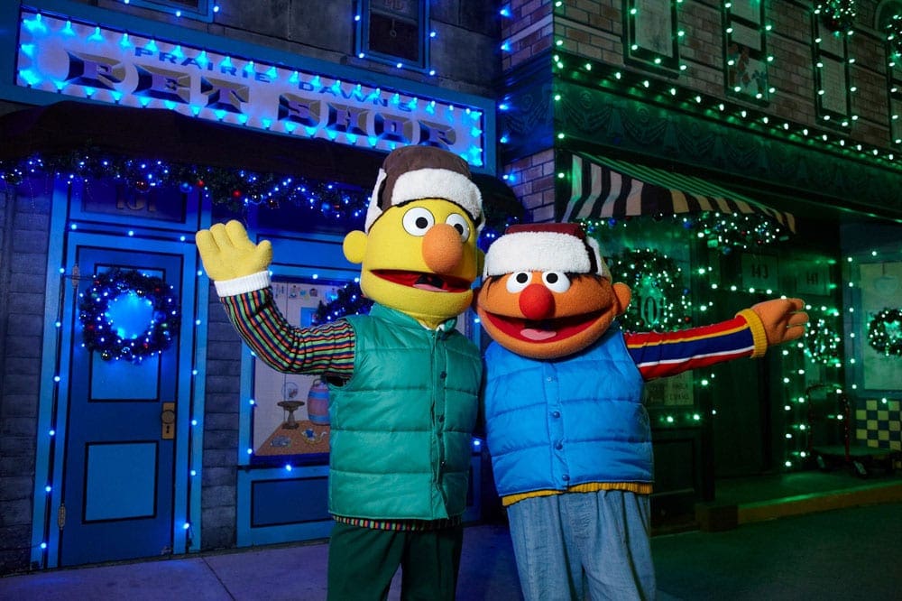 Burt and Ernie, wearing winter gear, stand arm and arm in front of blue and green Christmas lights.