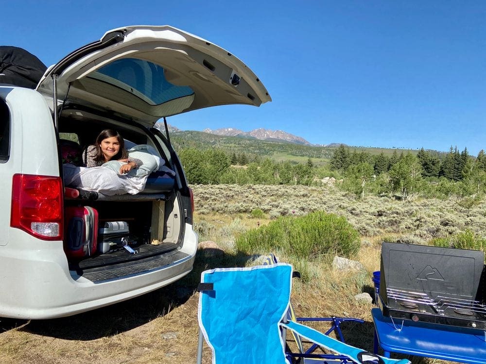 A young girl lays within a van while camping at the Blue River Campsite, nearby is their camping grill, , essential on our list of packing tips for COVID travel.