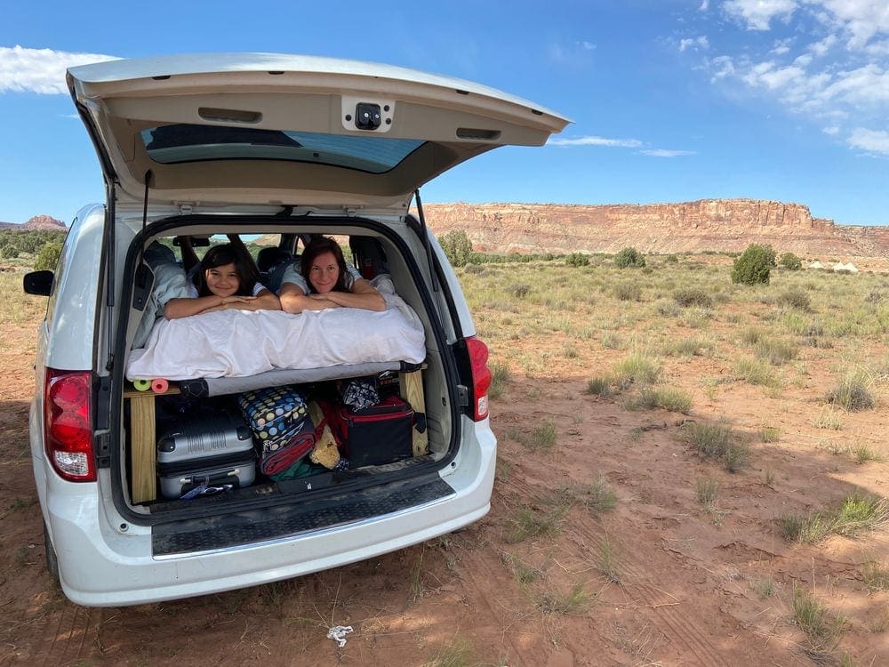 A mom and her daughter lie on a mattress within a van, while surrounded by sweeping natural views. Practical sleeping quarters are essential on our list of packing tips for COVID travel.