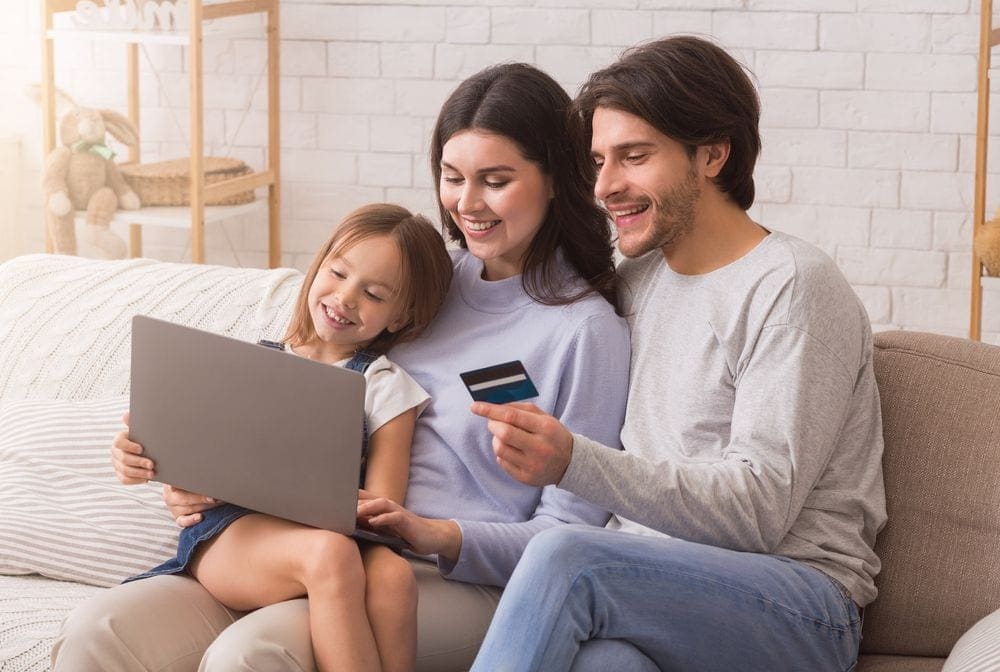 A man holds a credit card while sitting with his family looking at a computer, one of the things to remember in our Ultimate Ski Trip Packing List For Families This Winter.