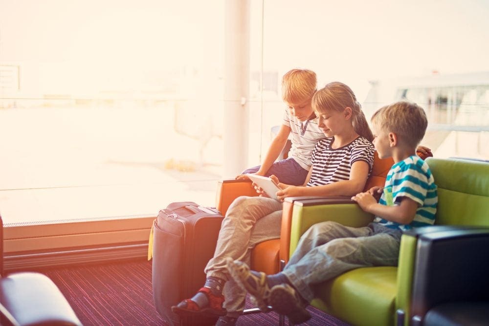 An older child holds a tablet while two younger children loon on in a waiting room at the airport after using a Travel Advisor for a Family Vacation.
