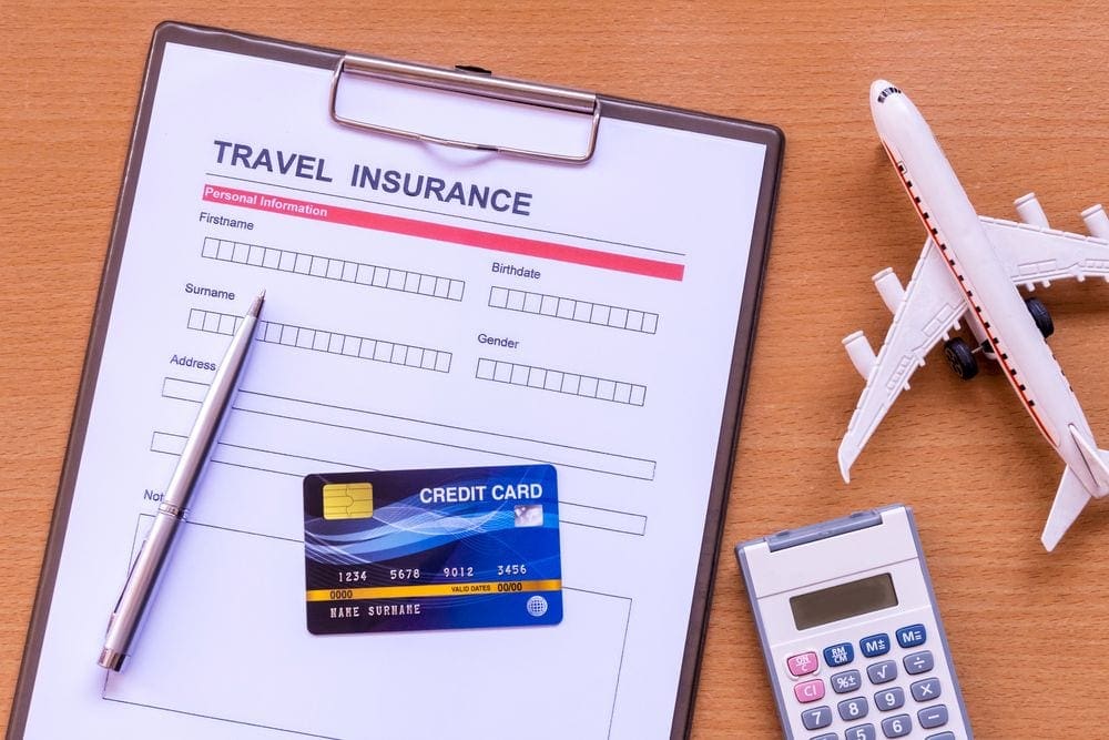 A clipboard holds a piece of paper reading "Travel Insurance", with a credit card and pen on top. An airplane and calculator rest nearby on the table. Added travel insurance is one factor families should consider when choosing a travel rewards card.