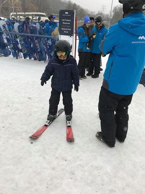 A young boy, dressed in a black snow suit, helmet, and ski googles, stands on skis next to a Mountain Creek ski instructor.