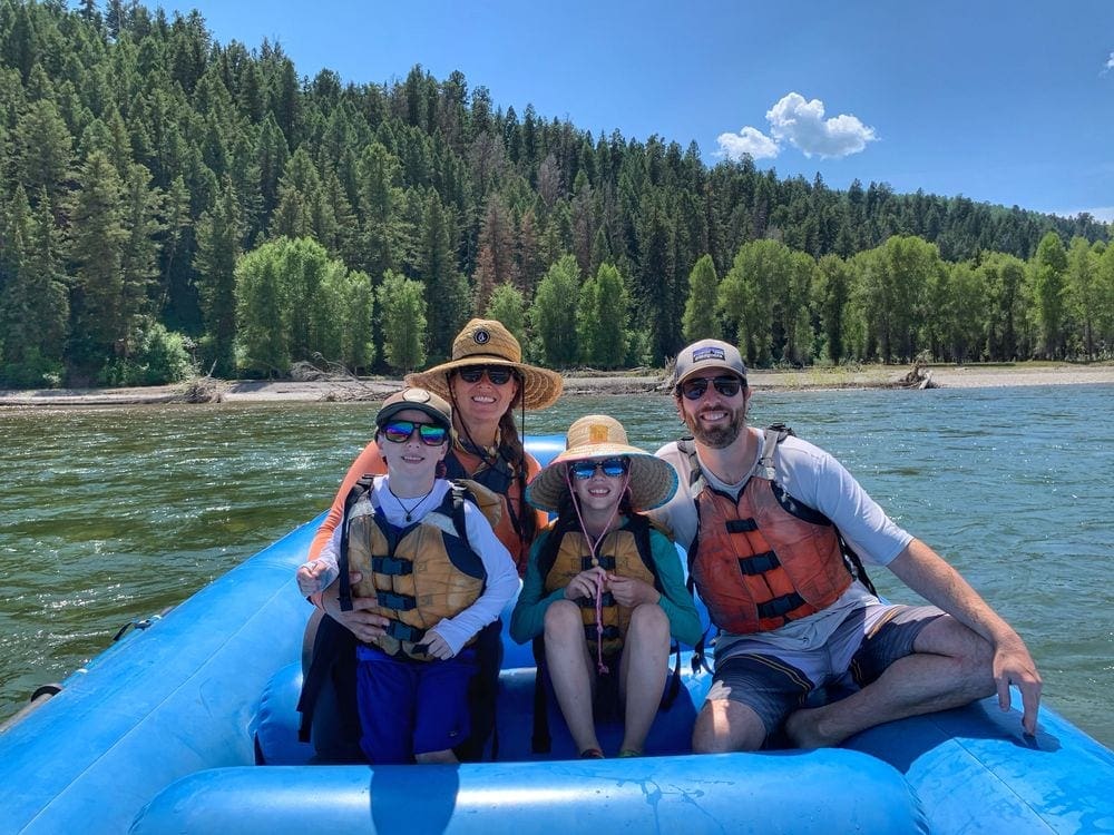A family of four huddles together, smiling, at the nose of a raft within a river, while a forest of evergreen trees sits on the shore.