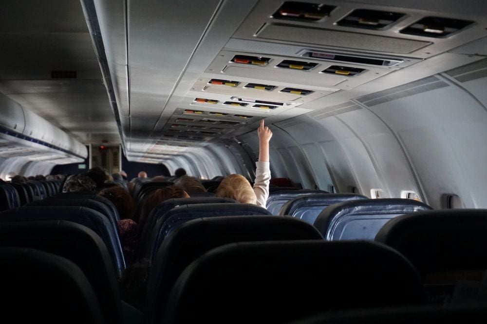 A young boy raises his hand to turn on the light inside an airplane cabin.