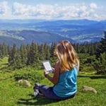 Girl holding a kindle and sitting top of a hill with a beautiful view of mountains