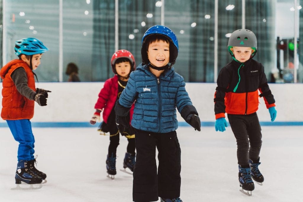 Several young boys wearing winter gear and helmets skate along the rink at Sky Rink at Chelsea Piers, one of the best indoor activities in New York City for kids.