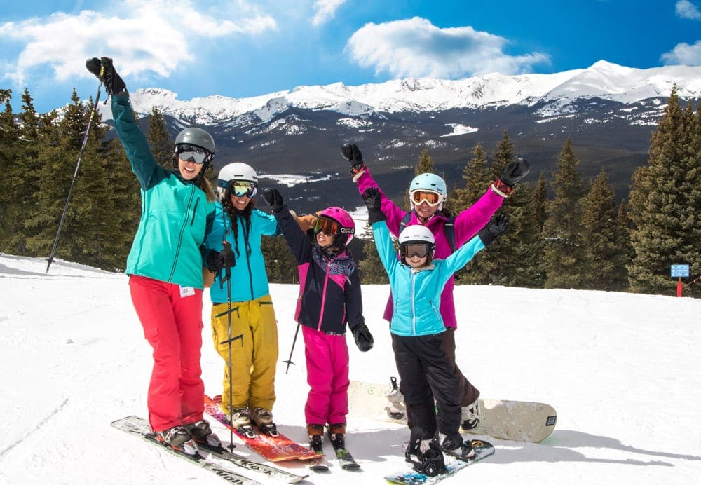 A family of five wearing colorful snow gear stands on skis while smiling at Cooper.