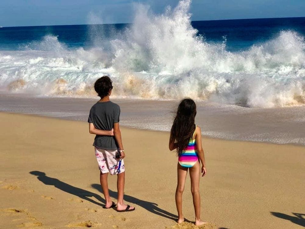 Two young kids stand on the beach in Los Cabos, while looking at crashing waves.