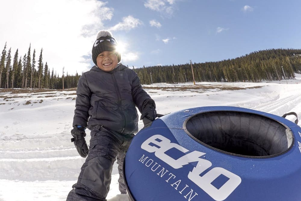 A young boy wearing a black snow suit sits in the snow holding onto a blue Echo Mountain snow tube.