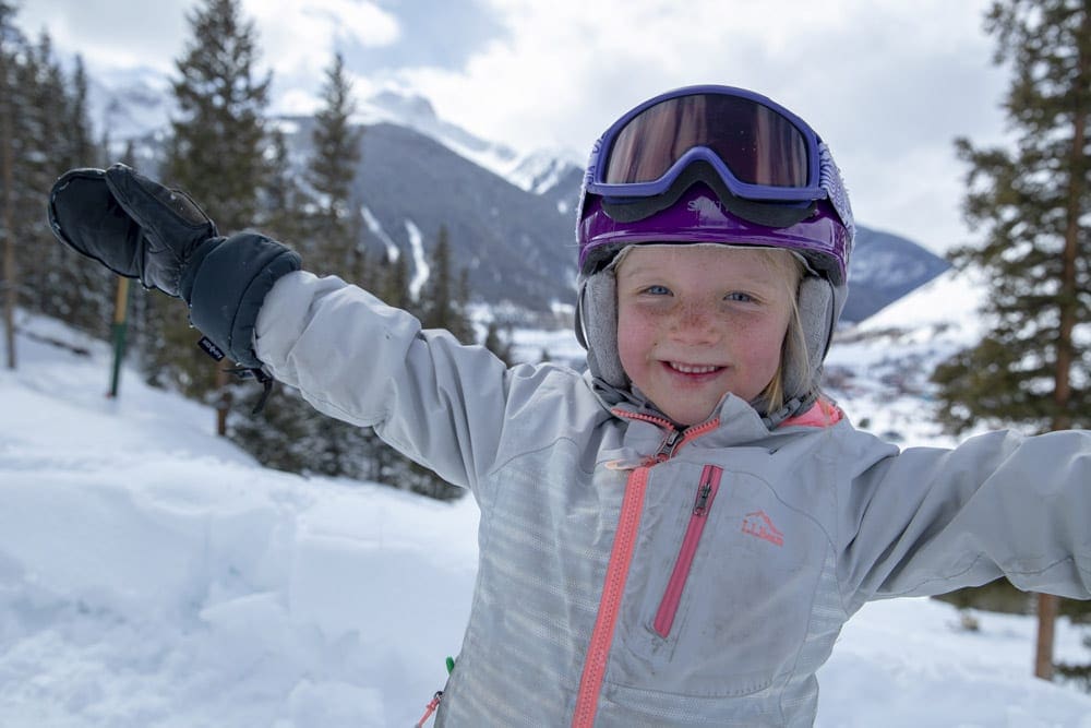 A young girl wearing a gray winter coat, helmet, and ski goggles holds her arms outstretched on Kendall Mountain.