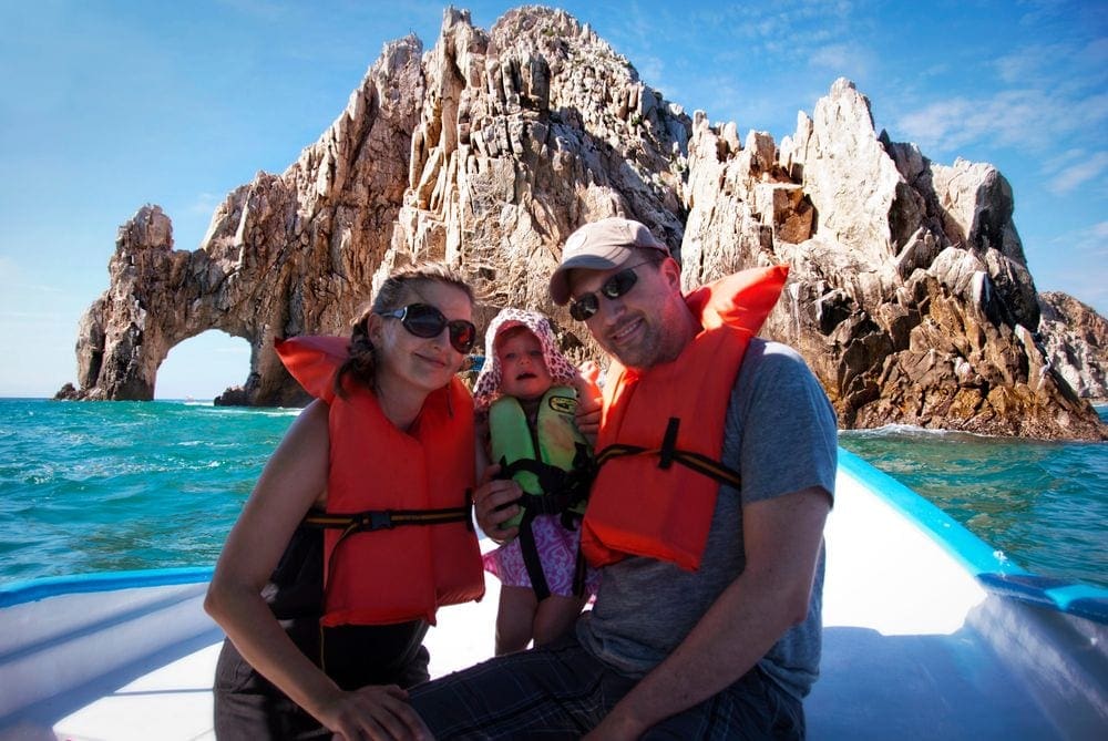 Parents sit at the front of a boat wearing orange life vests holding their infant daughter with the Lands End Arch in the background.