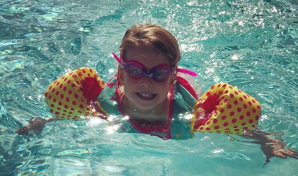 Little girl with blonde hair, pink goggles, and pink and yellow polkadot floats swimming in blue water.