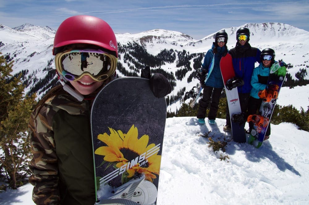 A young girl stands with a snowboard at Loveland Ski Area, one of the best small ski resorts in Colorado for families. Behind the young child stands three family members.